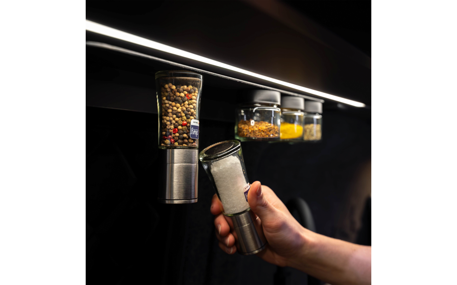 Hand removes a spice jar filled with coarse salt from a row of spice jars mounted on a rail under a light strip; three other jars with colorful spices visible