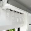 White overhead rack with five upside-down glasses: one short glass at left, four stemmed wine glasses to the right; a window with greenery is visible below