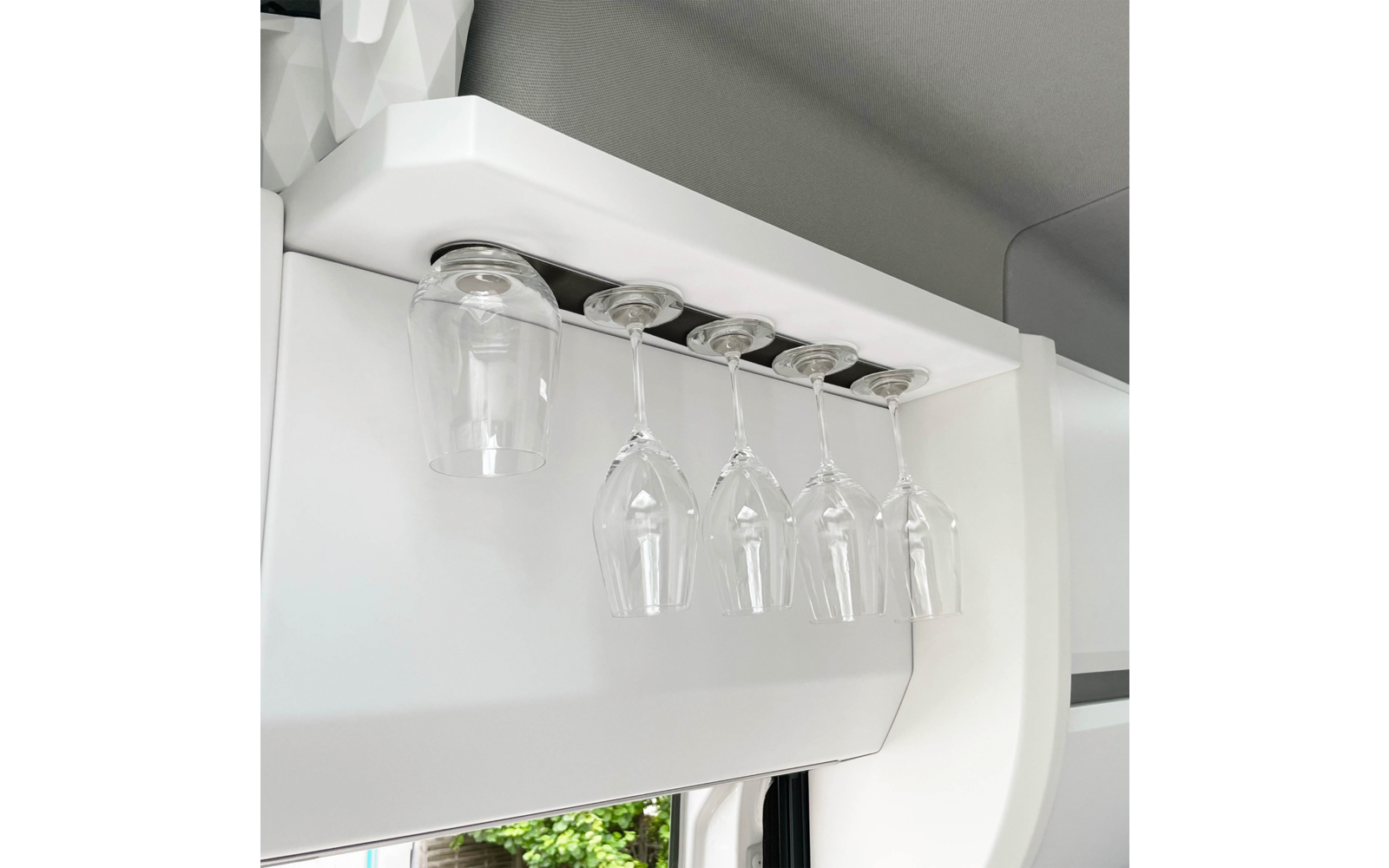 White overhead rack with five upside-down glasses: one short glass at left, four stemmed wine glasses to the right; a window with greenery is visible below