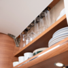 Open wooden kitchen cabinet with upside-down wine glasses on a rack, stacked plates, mugs and bowls