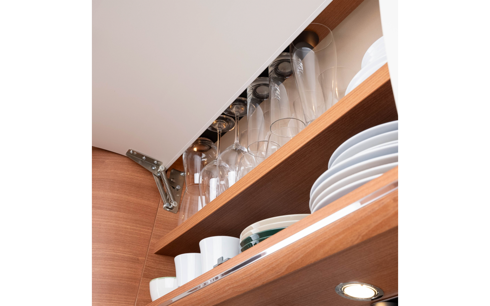 Open wooden kitchen cabinet with upside-down wine glasses on a rack, stacked plates, mugs and bowls