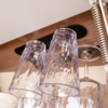 Several inverted faceted drinking glasses and one stemmed glass hanging from a wooden rack with circular cutouts; a metal spring is visible at the right