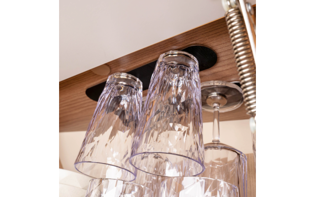 Several inverted faceted drinking glasses and one stemmed glass hanging from a wooden rack with circular cutouts; a metal spring is visible at the right