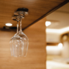 Two upside-down wine glasses hanging from a metal holder under a wooden shelf, blurred bright background