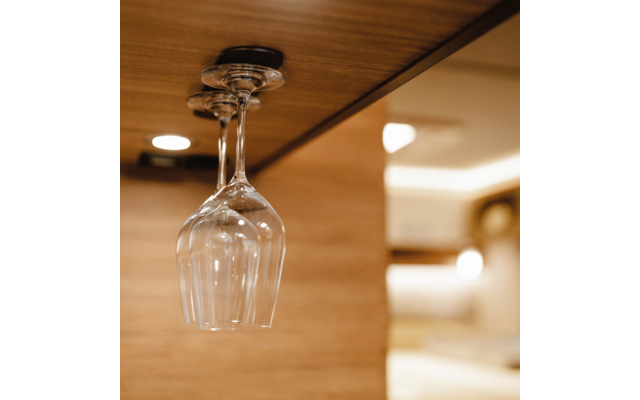 Two upside-down wine glasses hanging from a metal holder under a wooden shelf, blurred bright background