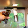 Hand holding two spice grinders with transparent containers (one with dark/multi-colored peppercorns, one with white salt) under a round recessed light beneath a wooden cabinet; background shows a window with green foliage and a faucet