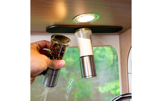 Hand holding two spice grinders with transparent containers (one with dark/multi-colored peppercorns, one with white salt) under a round recessed light beneath a wooden cabinet; background shows a window with green foliage and a faucet