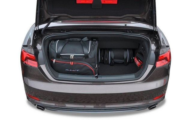Offener Kofferraum eines grauen Autos mit einer schwarzen Reisetasche mit roten Akzenten, zwei kleinen schwarzen Taschen und einem roten Objekt rechts