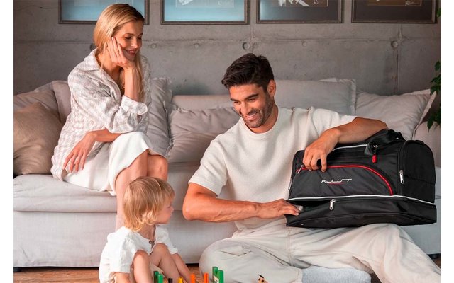 Mann sitzt auf dem Boden und öffnet eine schwarze Reisetasche, Frau sitzt auf dem Sofa, Kleinkind spielt mit bunten Bausteinen, Sofa mit Kissen und Bilder an der Wand