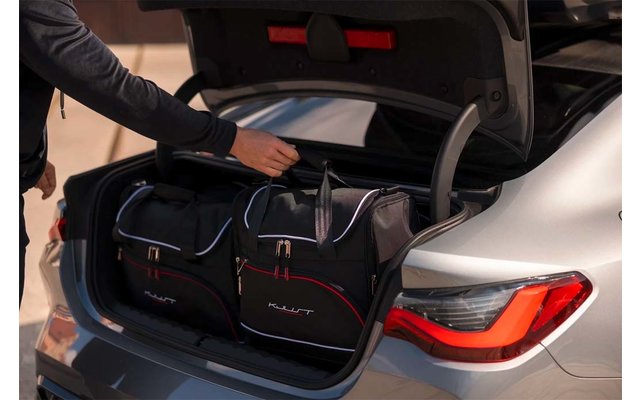 Geöffneter Kofferraum eines silbernen Autos mit zwei schwarzen Reisetaschen; eine Hand greift an den Henkel einer Tasche; rechte Heckleuchte sichtbar