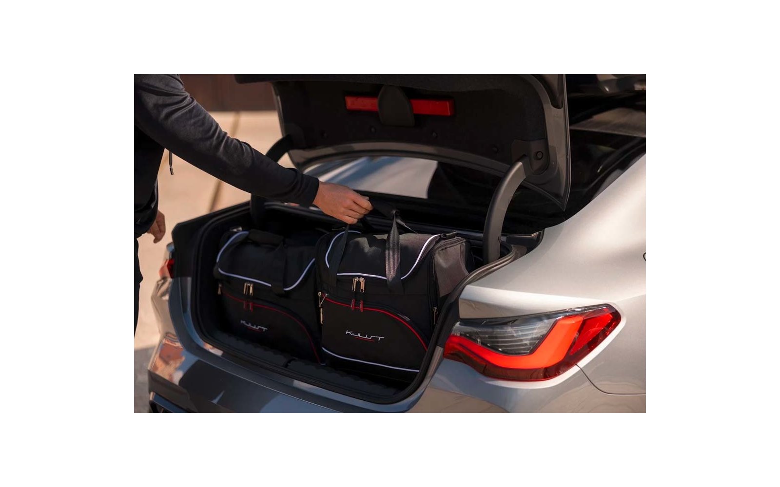 Geöffneter Kofferraum eines silbernen Autos mit zwei schwarzen Reisetaschen; eine Hand greift an den Henkel einer Tasche; rechte Heckleuchte sichtbar