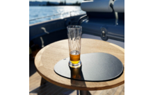 Half-filled beer glass on a round wooden table with a black placemat, water and part of a boat in the background