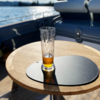 Half-filled beer glass on a round wooden table with a black placemat, water and part of a boat in the background