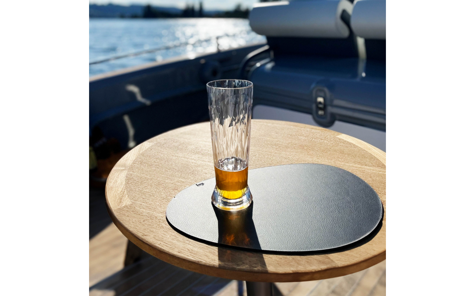 Half-filled beer glass on a round wooden table with a black placemat, water and part of a boat in the background