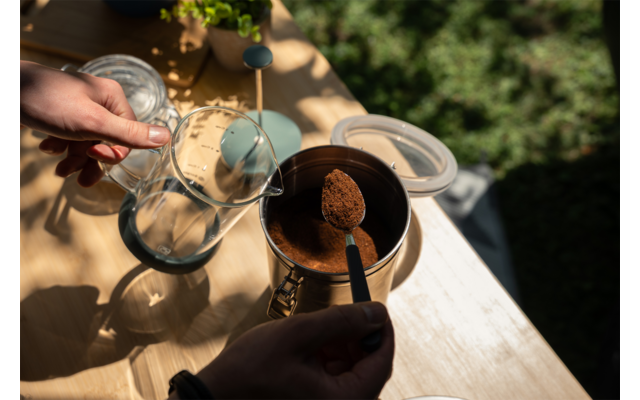 Two hands over a wooden table; one holds a glass carafe, the other a spoon with brown ground coffee above a metal container; on the table an open lid, a container with ground coffee and a small potted plant; blurred green background