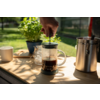 Hand pressing the plunger of a French press with coffee on a wooden table; nearby a cup, a glass jar, a stainless steel jug and a potted plant, garden in the background