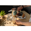 Two hands pouring coffee from a French press into a mug on a wooden table; croissant on a plate, knife, glass jar and small potted plant nearby