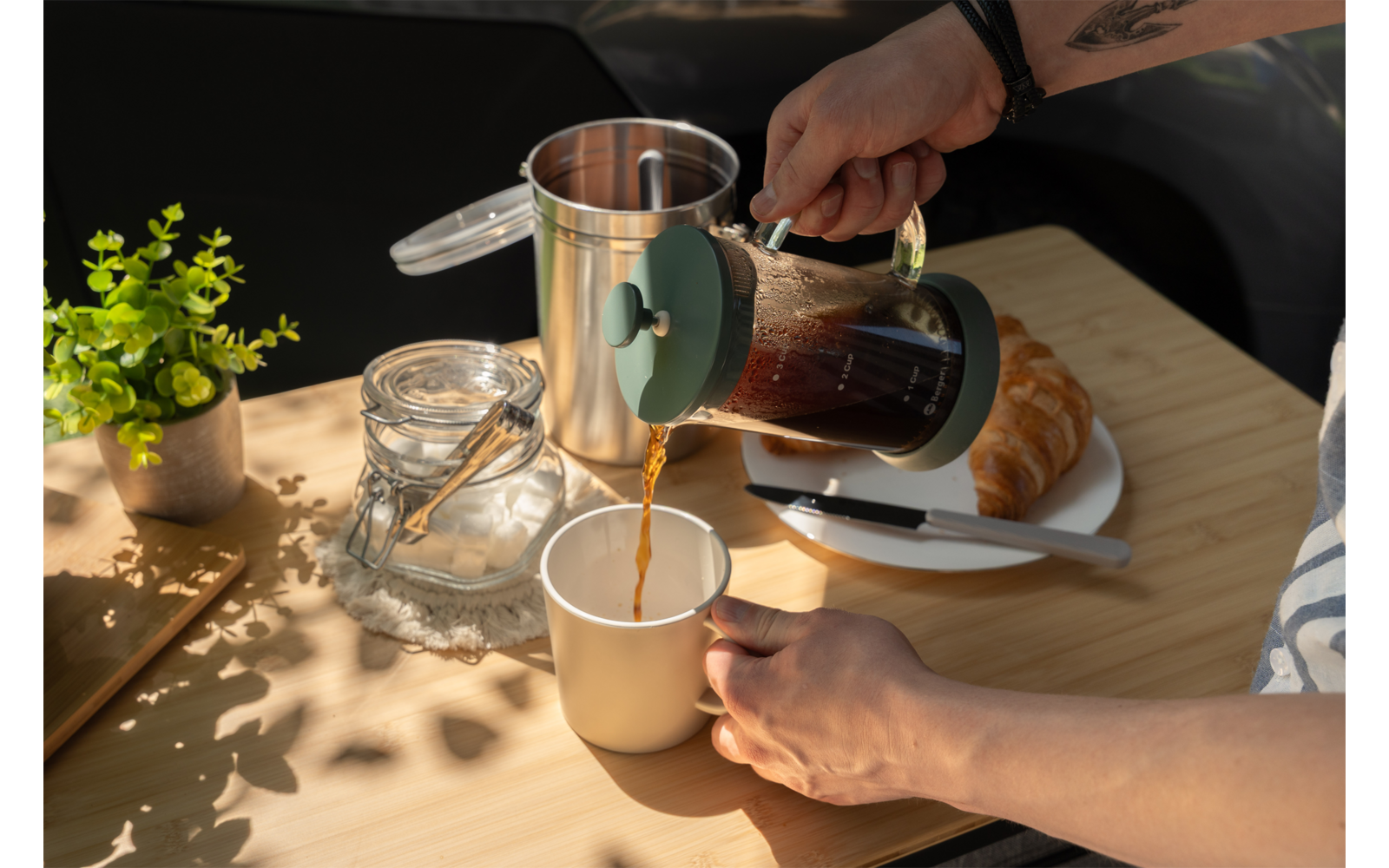 Two hands pouring coffee from a French press into a mug on a wooden table; croissant on a plate, knife, glass jar and small potted plant nearby