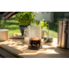 Glass carafe with measurement marks on a wooden surface; a kettle spout pours water into the carafe containing dark brown liquid; mug, small jar, metal canister and a potted green plant in the background, sunlight