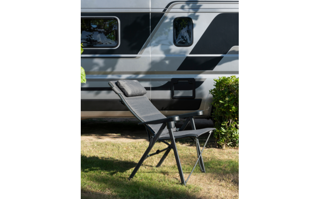 Klappliege mit Kopfkissen auf Gras vor einem silbernen Wohnwagen mit schwarzen Streifen, kleiner Busch und Baumschatten