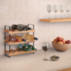 Kleines Holz-Weinregal mit schwarzem Metallgestell und sechs Weinflaschen auf einer Küchenarbeitsplatte; darüber an der Wand ein Regal mit vier leeren Weingläsern. Auf der Arbeitsplatte ein gefülltes Weinglas, eine Schale mit Obst und eine kleine Sch