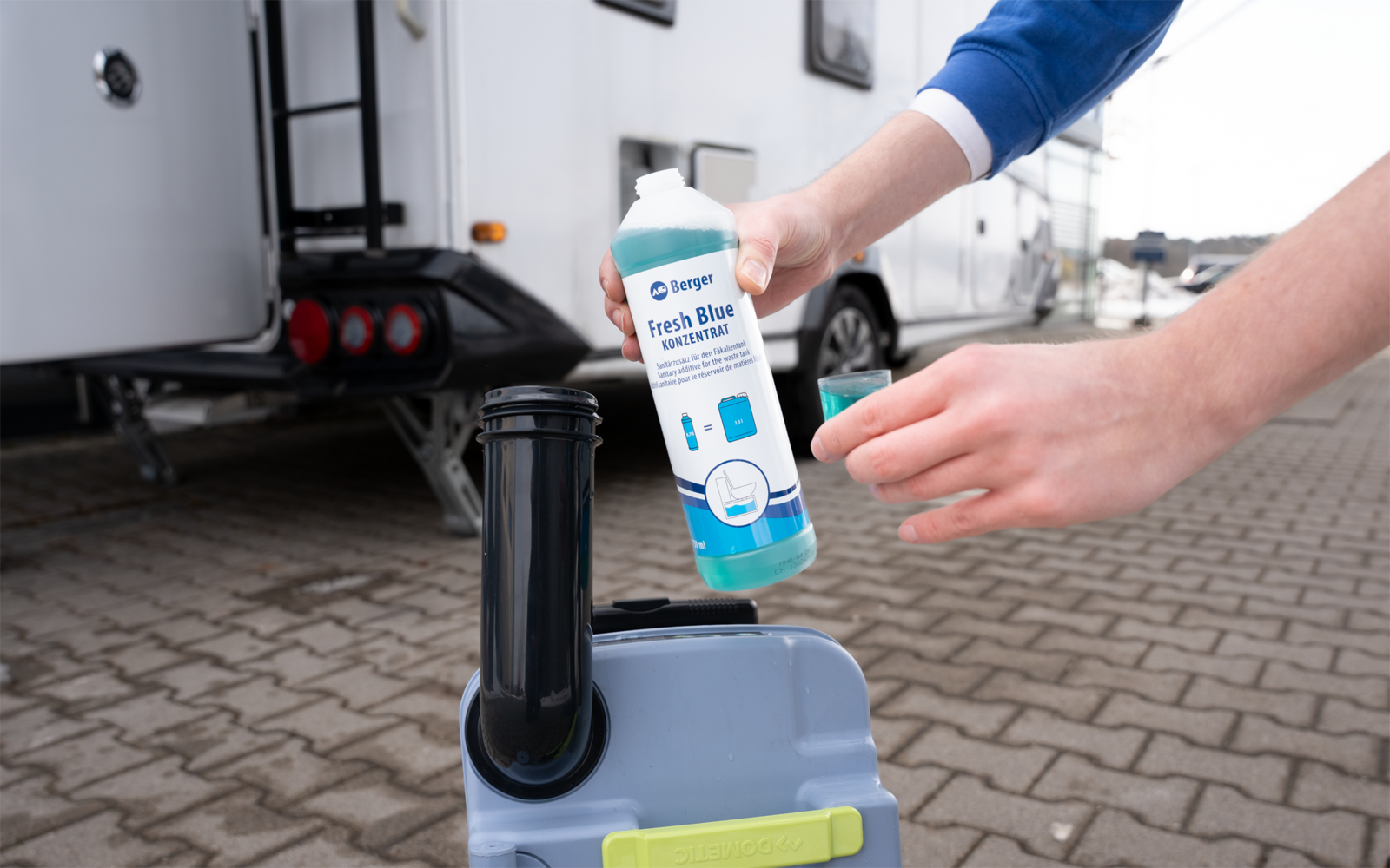 Zwei Hände halten eine Flasche mit blauem Etikett über einem grauen Kunststofftank mit schwarzem Einfüllrohr; im Hintergrund Wohnwagen auf gepflastertem Untergrund