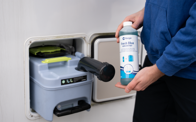 Person in blauem Pullover hält eine Flasche mit der Aufschrift 'Fresh Blue' und einen schwarzen Ausgussaufsatz über einer grauen Kassettentoilette mit grünem Deckel in einem offenen Fach