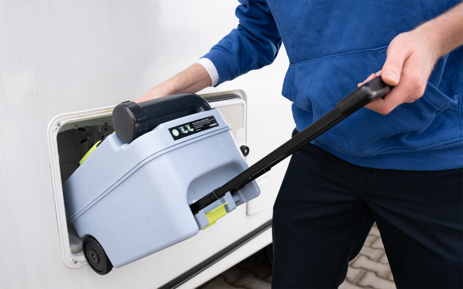 Person in blauem Oberteil zieht ein graues, rollbares Gerät mit schwarzem Griff und digitalem Display aus dem offenen Seitenfach eines weißen Wohnwagens; Pflasterboden sichtbar
