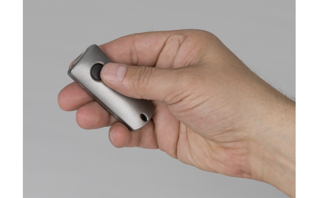 Rechte Hand, Daumen auf rundem schwarzen Knopf eines kleinen silbernen, abgerundeten rechteckigen Geräts vor neutralem grauem Hintergrund