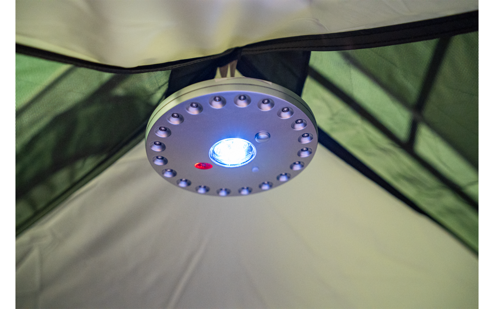Runde LED-Lampe befestigt an der Zeltdecke, mittig leuchtende blaue LED, umlaufende kleine LEDs, roter Knopf, grüner und beigefarbener Zeltstoff im Hintergrund
