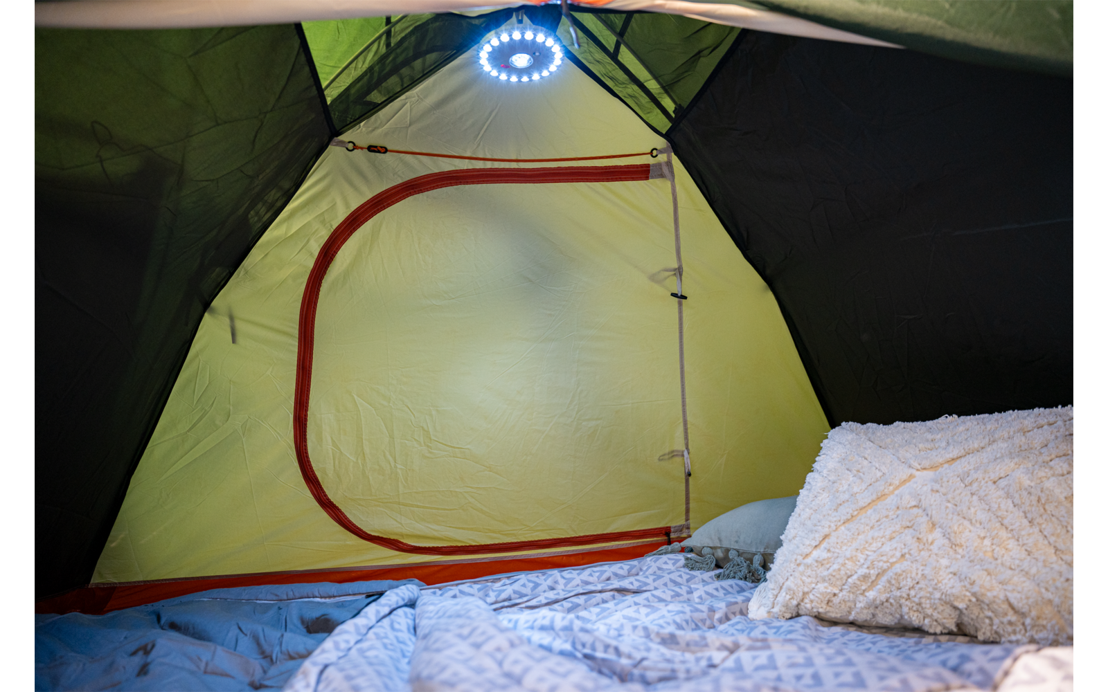 Innenraum eines Zeltes mit geschlossener Tür (roter Reißverschluss), hängender runder LED-Leuchte sowie Decke und Kissen auf dem Boden