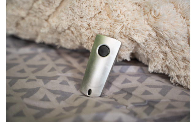 Kleines silbernes Gerät mit rundem schwarzen Knopf und kleinem Loch, lehnt an beigem flauschigem Kissen/Decke auf grauem, geometrisch gemustertem Bettbezug