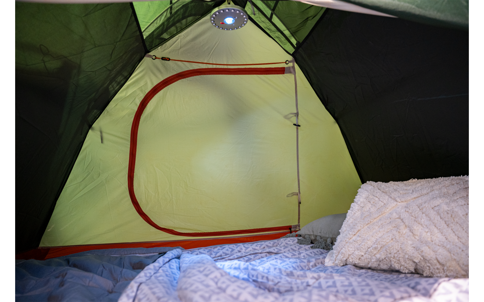 Innenraum eines grünen Zelts, Eingang mit rotem Reißverschluss, Lampe an der Decke, gemusterte Decke und strukturiertes Kissen auf einer Schlafunterlage