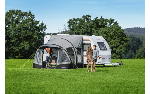 Caravan met aangebouwde voortent op een groen veld, twee personen staan voor de voortent, bomen en blauwe lucht op de achtergrond