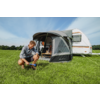 Man knielt op het gras en spant een spanlijn van de voortent voor een caravan; vrouw zit aan een tafel in de voortent; groen veld, blauwe lucht