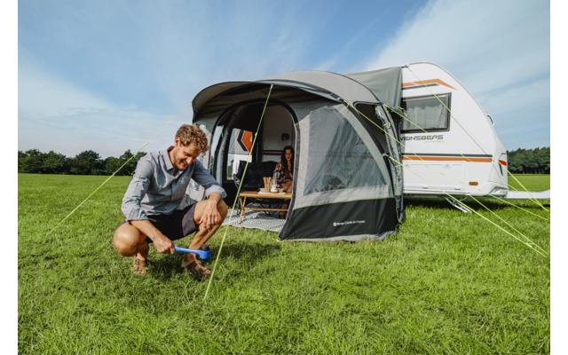 Man knielt op het gras en spant een spanlijn van de voortent voor een caravan; vrouw zit aan een tafel in de voortent; groen veld, blauwe lucht