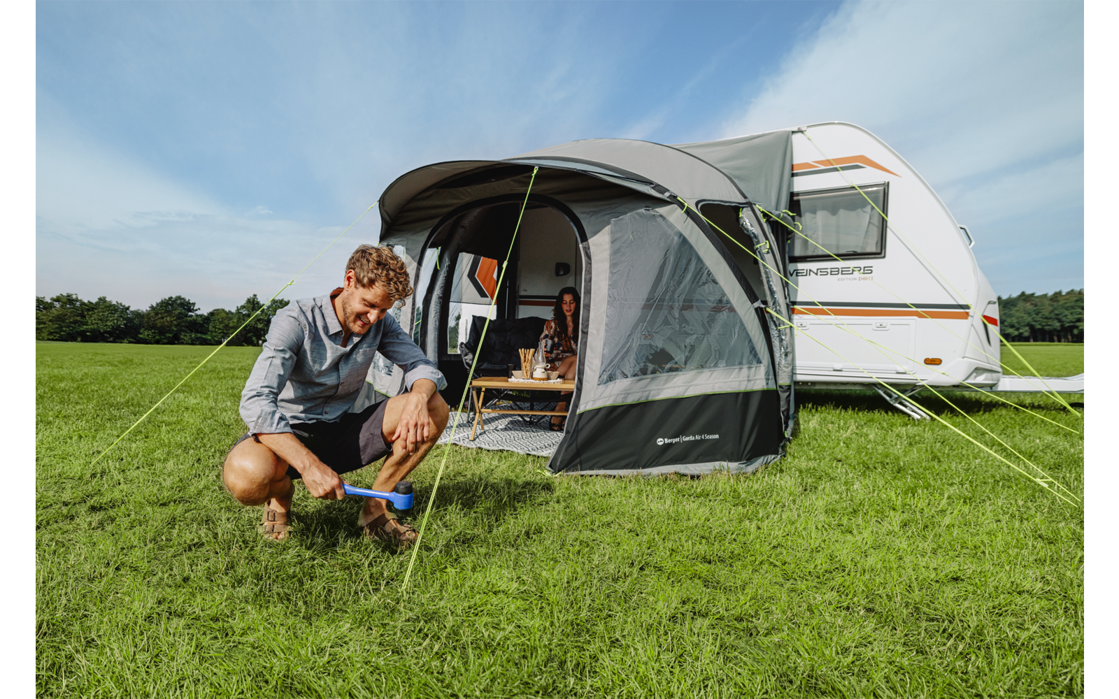 Man knielt op het gras en spant een spanlijn van de voortent voor een caravan; vrouw zit aan een tafel in de voortent; groen veld, blauwe lucht