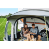 Een man en een vrouw zitten op vouwstoelen in een voortent naast een caravan, ze houden bekers vast; voor hen staat een klein tafeltje met bekers en voorwerpen; grasveld en bomen op de achtergrond, caravanwiel zichtbaar