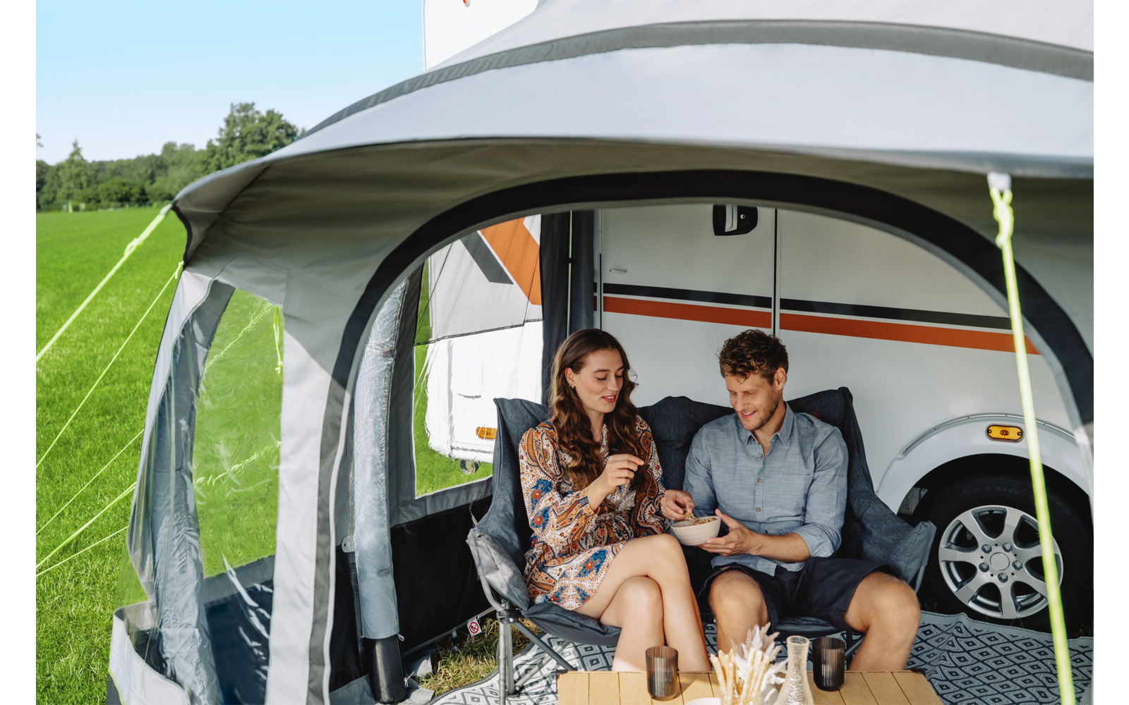 Een man en een vrouw zitten op vouwstoelen in een voortent naast een caravan, ze houden bekers vast; voor hen staat een klein tafeltje met bekers en voorwerpen; grasveld en bomen op de achtergrond, caravanwiel zichtbaar