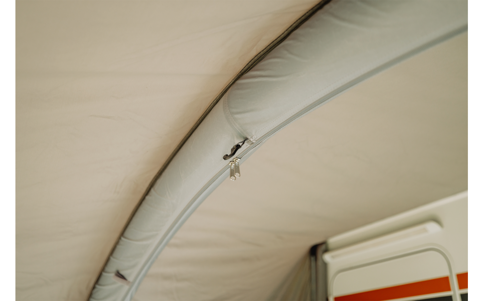 Binnenaanzicht van een stoffen dak met een gebogen, gestikte buis en een open rits met een klein metalen ritstrekker; rechts zichtbaar: een wit paneel met een oranje streep