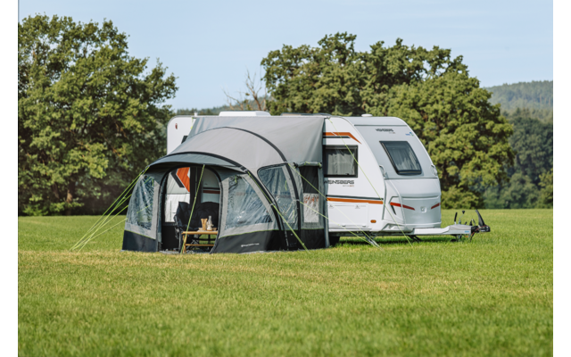 Witte caravan met aangebouwd voortent op een groen weiland, bomen op de achtergrond