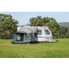 Witte caravan met grijze voortent op een groen veld, bomen en blauwe lucht op de achtergrond