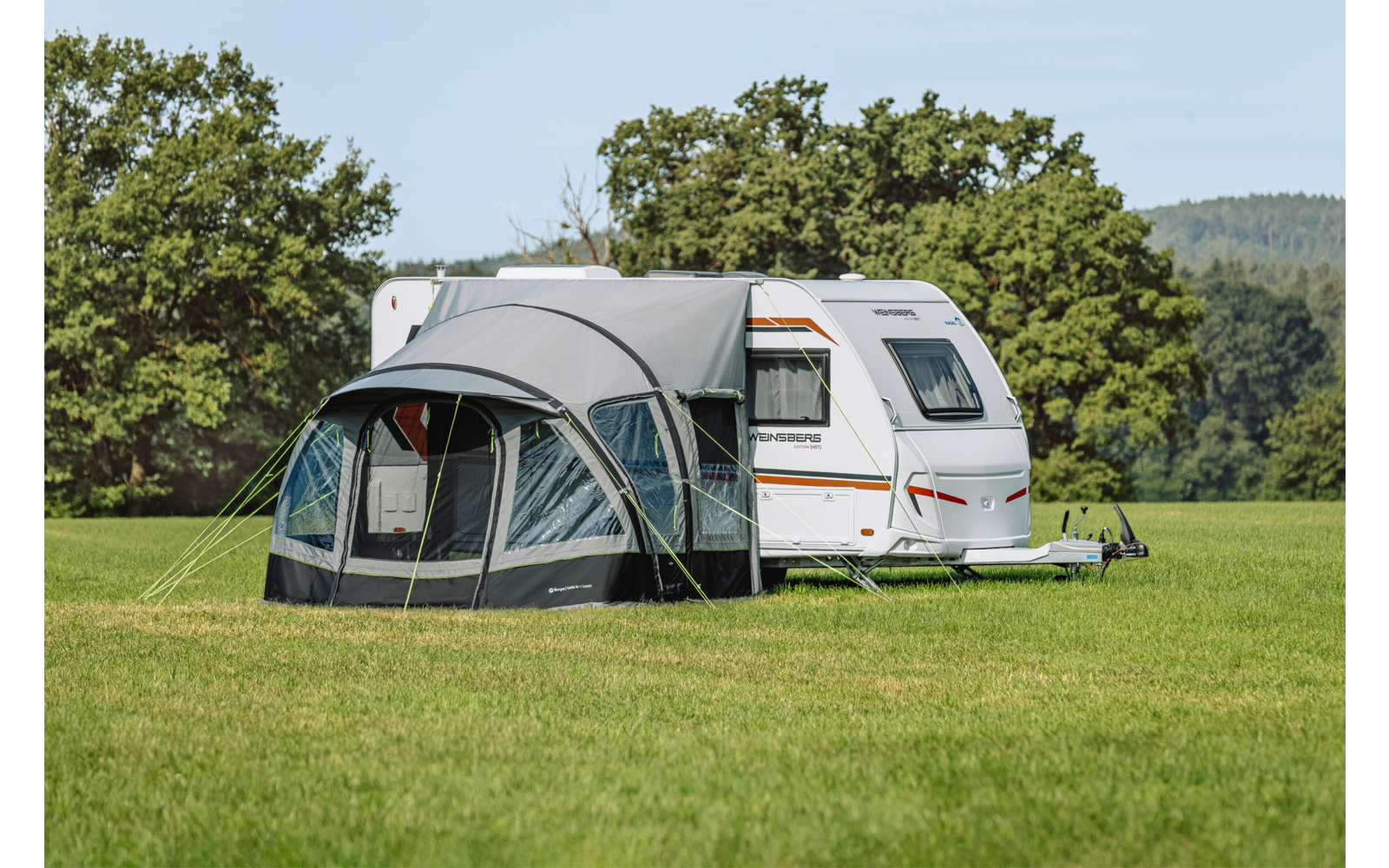 Witte caravan met grijze voortent op een groen veld, bomen en blauwe lucht op de achtergrond