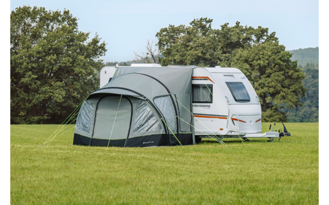 Witte caravan met grijs voortent op een groen veld, bomen op de achtergrond
