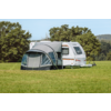 Witte caravan op een groen veld met een grijze voortent bevestigd, bomen op de achtergrond en blauwe lucht