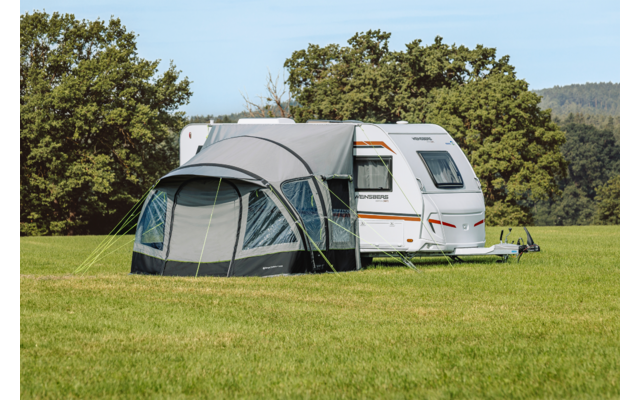 Witte caravan op een groen veld met een grijze voortent bevestigd, bomen op de achtergrond en blauwe lucht