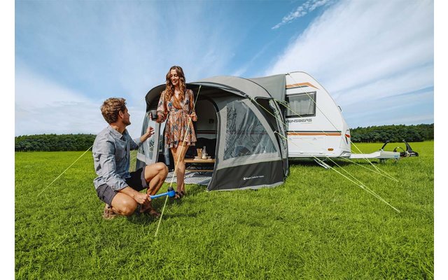 Een man knielt op het gras en geeft een beker aan een vrouw voor een caravan met een grijze voortent; groen gras en blauwe lucht met wolken
