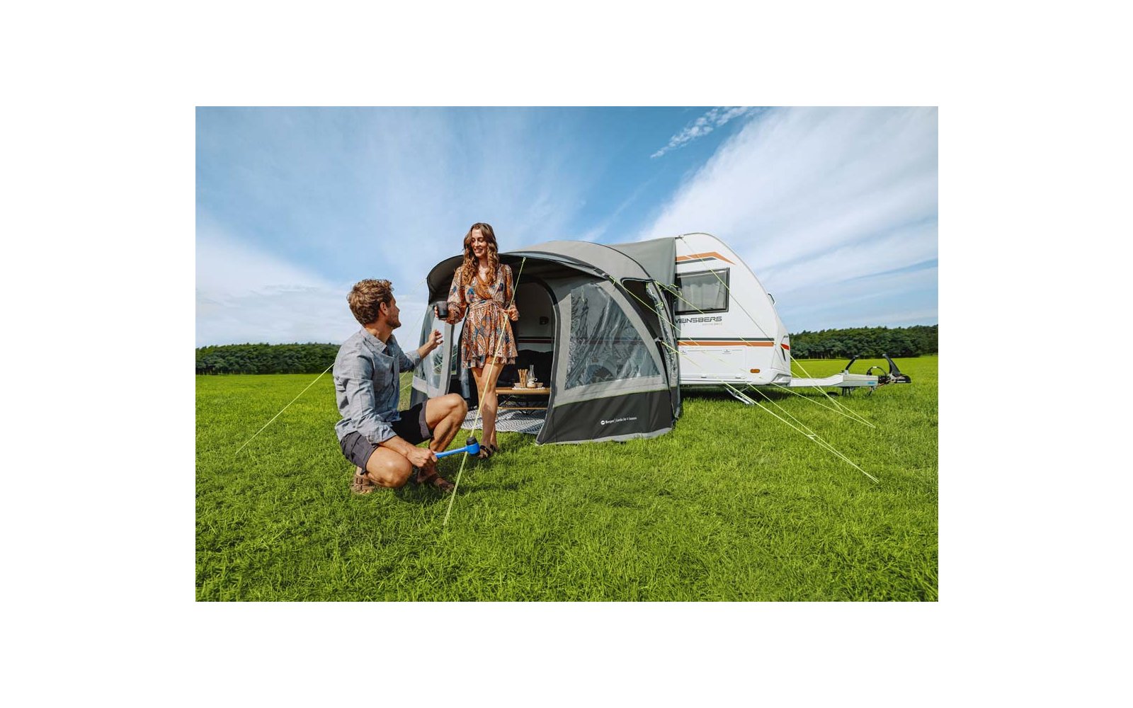 Een man knielt op het gras en geeft een beker aan een vrouw voor een caravan met een grijze voortent; groen gras en blauwe lucht met wolken