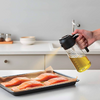 Hand hält Glas-Ölspender mit schwarzem Ausguss und gießt dünnen Ölstrahl auf vier Lachsfilets in einem mit Backpapier ausgelegten Backblech; Arbeitsplatte, Schalen und Wasserhahn im Hintergrund