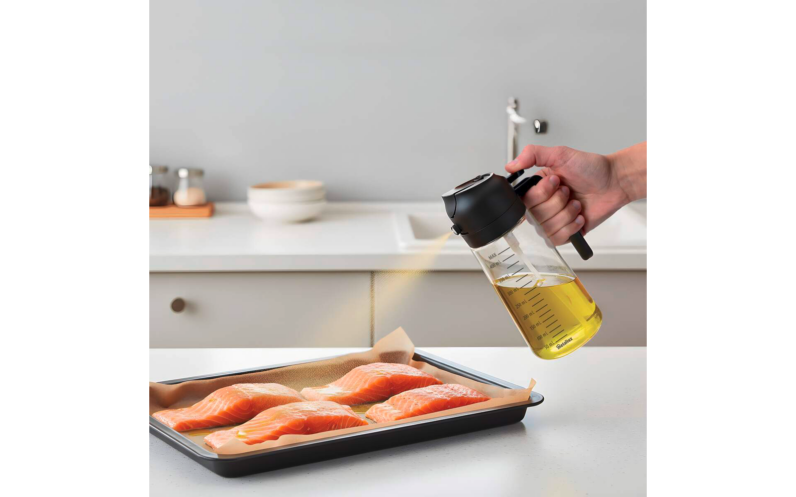 Hand hält Glas-Ölspender mit schwarzem Ausguss und gießt dünnen Ölstrahl auf vier Lachsfilets in einem mit Backpapier ausgelegten Backblech; Arbeitsplatte, Schalen und Wasserhahn im Hintergrund